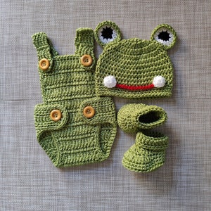 May include: A handmade, green crochet baby outfit set, including a romper with wooden buttons, a frog hat with white pom-poms and black and white eyes, and matching booties. The set is designed to resemble a frog.