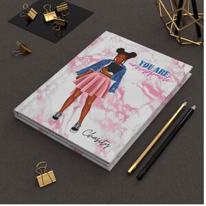 May include: A hardbound journal with a marble design and an illustration of a person wearing a denim jacket and pink skirt. The text "YOU ARE unstoppable" is printed on the cover, along with the name "Charity". Gold and black pencils and binder clips are also visible.