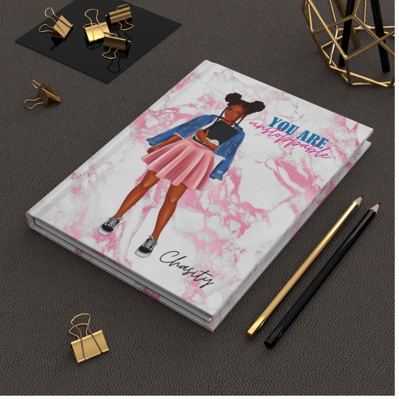 May include: A hardbound journal with a marble design and an illustration of a person wearing a denim jacket and pink skirt. The text "YOU ARE unstoppable" is printed on the cover, along with the name "Charity". Gold and black pencils and binder clips are also visible.