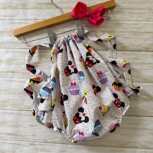 May include: A white baby romper with a colorful pattern of cartoon characters and cupcakes. The romper is hanging on a wooden hanger with a bright pink bow attached.