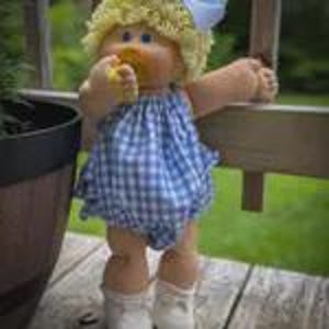 May include: A doll with curly blonde yarn hair and blue eyes, wearing a blue and white gingham romper with a ruffled neckline. The doll has a tan complexion, a pacifier in its mouth, and is wearing white shoes with white socks.