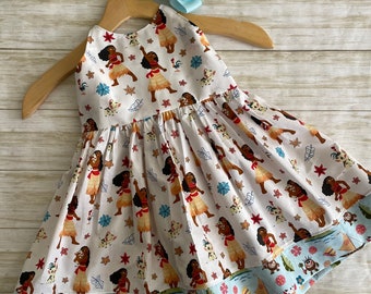 May include: A white sleeveless dress with a blue and green trim. The dress features a repeating pattern of a cartoon character with brown skin, black hair, and a red skirt. The character is holding a pig and is surrounded by flowers and leaves.