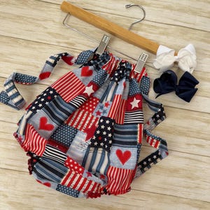 May include: A patriotic baby outfit with a patchwork design featuring red, white, and blue patterns, including stars, stripes, and hearts. The outfit is displayed on a wooden hanger, with two decorative bows in white and navy blue.