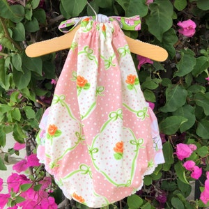 May include: A pink and white baby onesie with a floral print and green bows. The onesie has a ruffled hem and is hanging on a wooden hanger.
