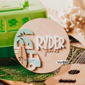May include: A round wooden sign with the name "RYDER" in white 3D letters, and the name "james" in a script font. The sign features a surfboard, palm trees, and a camper van in pastel colors. The Wild Born logo is visible.