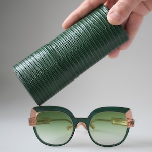 Könnte beinhalten: Grün-goldene Sonnenbrille mit Farbverlaufsgläsern, kombiniert mit einem passenden grünen, strukturierten Etui. Die Sonnenbrille hat ein einzigartiges Design mit goldenen Akzenten und einem hellgrünen Farbton. Das Etui wird über der Brille gehalten.