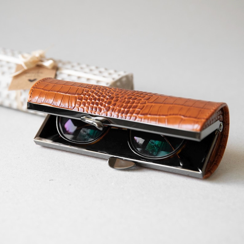 K&ouml;nnte beinhalten: Ein braunes Brillenetui mit Krokodiltextur und silbernem Rahmen, ge&ouml;ffnet, um eine runde Sonnenbrille zu zeigen. Im Hintergrund befindet sich eine Geschenkbox mit Schleife.