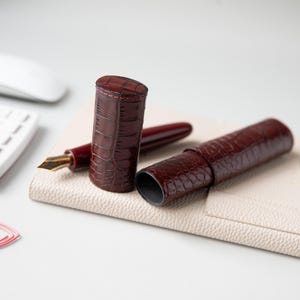 May include: A dark burgundy fountain pen with a gold-toned nib and a matching two-piece cylindrical pen case. Both the pen and the case feature a glossy, embossed crocodile skin texture with visible stitching along the edges of the case.