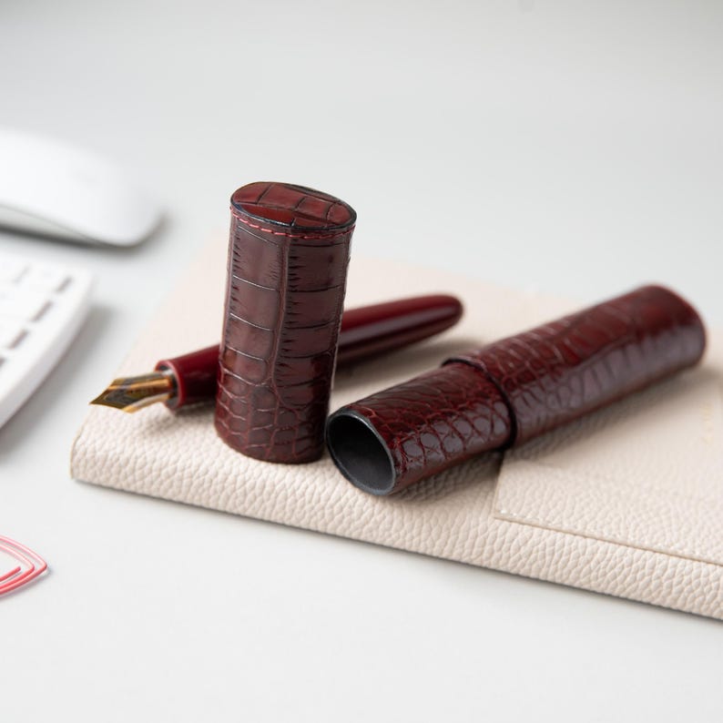 May include: A dark burgundy fountain pen with a gold-toned nib and a matching two-piece cylindrical pen case. Both the pen and the case feature a glossy, embossed crocodile skin texture with visible stitching along the edges of the case.