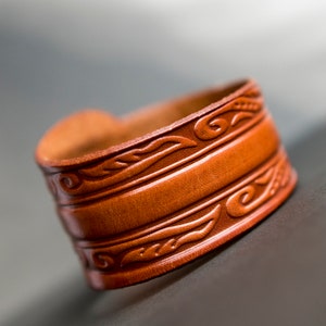 May include: A brown leather cuff bracelet with intricate floral and swirl designs. The bracelet is made of genuine leather and has a smooth, polished finish.