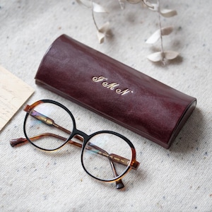 May include: A pair of round, tortoiseshell eyeglasses with black frames and clear lenses. The glasses are resting on a white, textured surface next to a burgundy leather glasses case with the initials "J.M.N" embossed in gold.