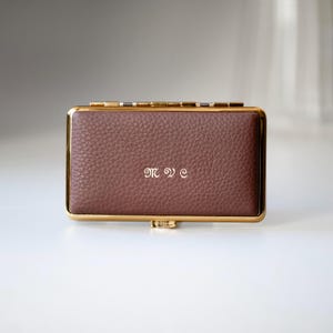 Puede incluir: Un estuche rectangular de cuero marrón con ribetes dorados y un cierre. El estuche tiene las iniciales "M W E" en una delicada escritura plateada en la parte delantera. El estuche está cerrado y sobre una superficie blanca.