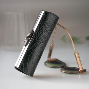 K&ouml;nnte beinhalten: Ein dunkelgr&uuml;nes, strukturiertes Brillenetui mit silbernem Verschluss und Zierleiste. Das Etui befindet sich neben einer Brille mit goldfarbenem Gestell und runden Gl&auml;sern. Das Etui hat ein Krokodilleder-Muster.