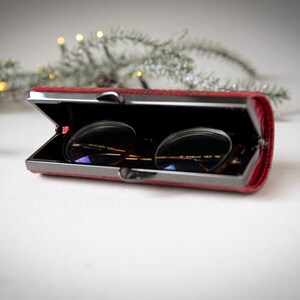 K&ouml;nnte beinhalten: Ein rotes, rechteckiges Brillenetui mit Metallverschluss, ge&ouml;ffnet, um eine Brille zu zeigen. Das Etui hat eine strukturierte Oberfl&auml;che und ein schwarzes Innenfutter. Die Brille hat dunkle Gl&auml;ser und einen d&uuml;nnen Metallrahmen.
