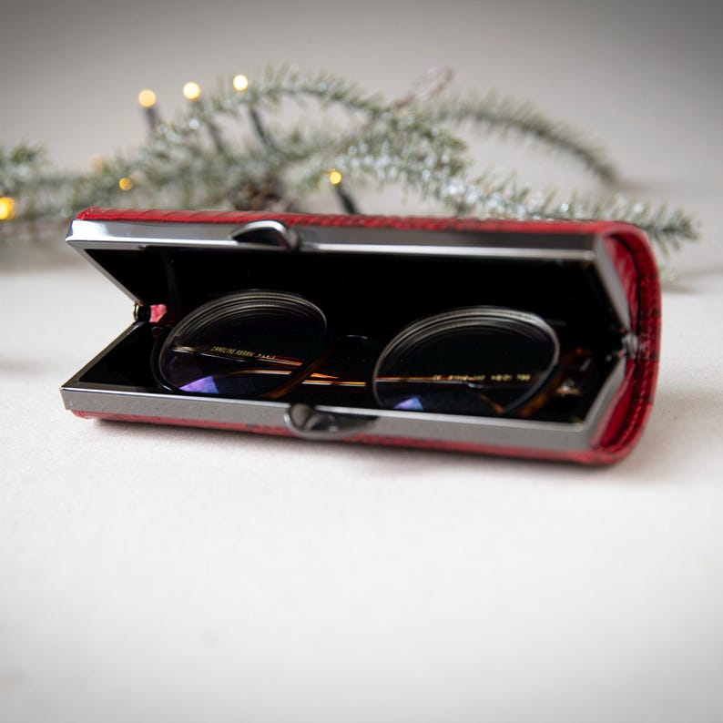 K&ouml;nnte beinhalten: Ein rotes, rechteckiges Brillenetui mit Metallverschluss, ge&ouml;ffnet, um eine Brille zu zeigen. Das Etui hat eine strukturierte Oberfl&auml;che und ein schwarzes Innenfutter. Die Brille hat dunkle Gl&auml;ser und einen d&uuml;nnen Metallrahmen.