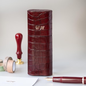 Op de afbeelding: Een bordeauxrode leren pennenkoker met een krokodillenstructuur en gouden initialen "WH". Een bordeauxrode vulpen, een rode lakzegel en decoratieve tapijtrollen zijn ook zichtbaar. De koker staat rechtop.