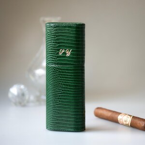 May include: A green, textured leather cigar case with gold initials "S.Y." on the front. A lit cigar rests on a white surface next to the case. The case is cylindrical and has a reptile-like pattern.