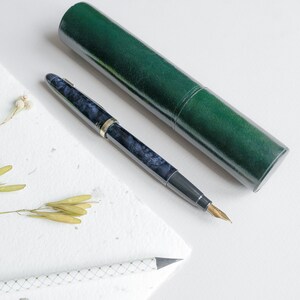 May include: A dark blue and silver fountain pen with a gold nib, resting on a white surface next to a green pen case.