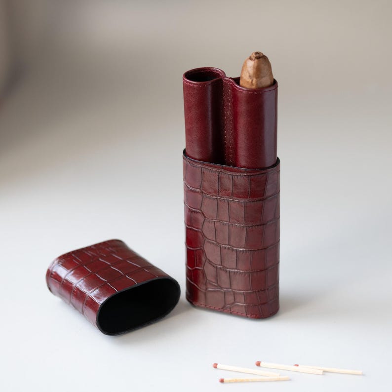 May include: A burgundy leather cigar case with a crocodile texture, holding a cigar. The case is open, with the lid lying beside it. Three matches are visible. The case is designed to hold two cigars.