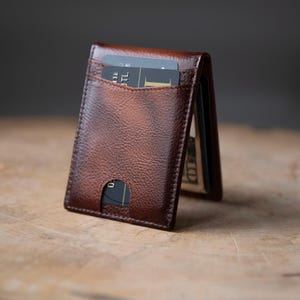 Op de afbeelding: Een donkerbruine lederen kaarthouder met een getextureerde afwerking. De portemonnee heeft een verticaal vak met een zwarte creditcard met zichtbare tekst en een zijvak voor papiergeld. Het ontwerp bevat een uitsparing voor de duim.