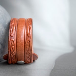 May include: A brown leather bracelet with an intricate embossed design. The bracelet has a silver buckle.