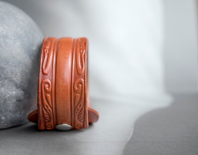 May include: A brown leather bracelet with an intricate embossed design. The bracelet has a silver buckle.