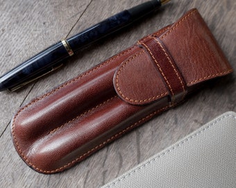 May include: A brown leather pen case with two slots for pens. The case has a flap that closes with a snap. A blue and black fountain pen is lying next to the case.