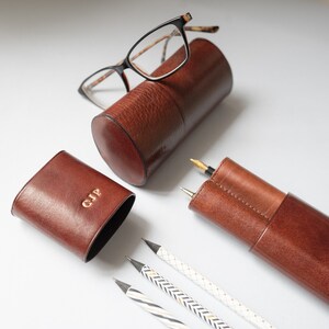 K&ouml;nnte beinhalten: Zwei braune Lederetuis, eines zylindrisch und eines rechteckig, mit goldenen Lettern. Das zylindrische Etui enth&auml;lt eine Brille mit schwarzen Gl&auml;sern. Das rechteckige Etui hat die Initialen "GJP" auf der Vorderseite gepr&auml;gt. Drei Bleistifte mit wei&szlig;en und goldenen Streifen liegen auf einer wei&szlig;en Oberfl&auml;che.