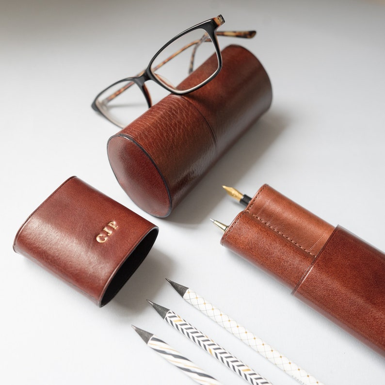 K&ouml;nnte beinhalten: Zwei braune Lederetuis, eines zylindrisch und eines rechteckig, mit goldenen Lettern. Das zylindrische Etui enth&auml;lt eine Brille mit schwarzen Gl&auml;sern. Das rechteckige Etui hat die Initialen "GJP" auf der Vorderseite gepr&auml;gt. Drei Bleistifte mit wei&szlig;en und goldenen Streifen liegen auf einer wei&szlig;en Oberfl&auml;che.