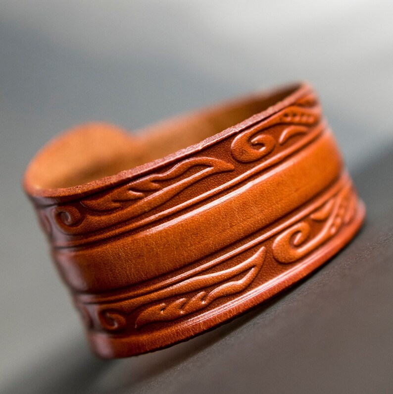 May include: A brown leather bracelet with intricate embossed designs. The bracelet has a wide band and is decorated with a repeating pattern of leaves and swirls.