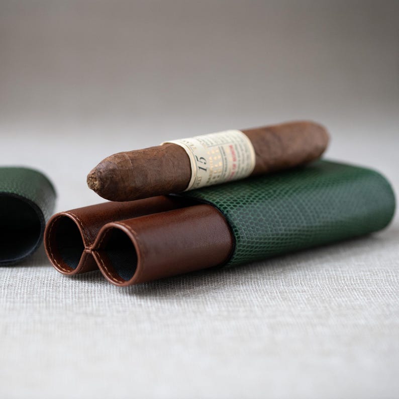 May include: A brown cigar with a white label rests on a green and brown leather cigar case. The case has a textured finish and a dual-tube design. The cigar is a dark brown color.