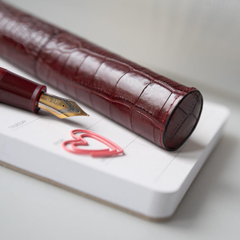 May include: A deep burgundy fountain pen with a gold-toned nib and a matching cylindrical pen case featuring a textured crocodile-embossed pattern. A small pink heart-shaped paperclip rests on a white planner page next to the writing instrument.