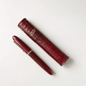 Op de afbeelding: Een bordeauxrode vulpen met gouden accenten en een bijpassende leren etui. De pennenzak heeft een krokodillenleertextuur en gouden initialen. De pen ligt naast de etui op een witte ondergrond.