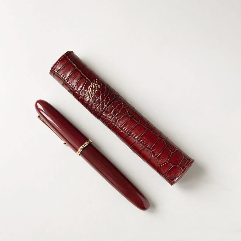 May include: A deep burgundy fountain pen with a gold-colored band and a matching cylindrical pen case featuring a textured crocodile-skin pattern and gold-colored monogrammed initials embossed on the surface of the dark red material.
