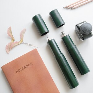 May include: A green leather notebook with the word "NOTEBOOK" embossed on the cover.  The notebook is surrounded by various writing implements, including a green leather pen case, a green leather pencil case, and a bottle of ink.