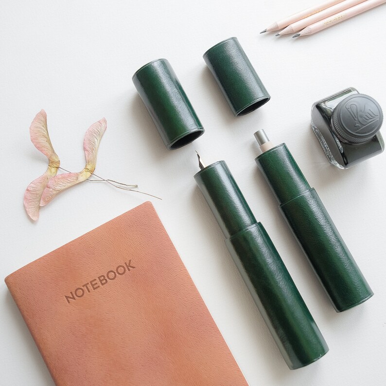 May include: A green leather notebook with the word "NOTEBOOK" embossed on the cover.  The notebook is surrounded by various writing implements, including a green leather pen case, a green leather pencil case, and a bottle of ink.