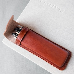 Può includere: Un astuccio per penne in pelle marrone con chiusura a zip. L'astuccio è appoggiato su una superficie bianca con il testo "PLANNING" in rilievo.