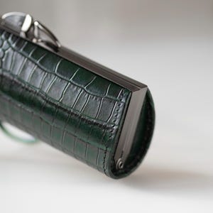 K&ouml;nnte beinhalten: Eine dunkelgr&uuml;ne, rechteckige Clutch mit Krokodilhautstruktur. Die Clutch hat einen dunklen Metallrahmen und -verschluss. Die Tasche hat eine zylindrische Form und scheint aus Leder zu sein.