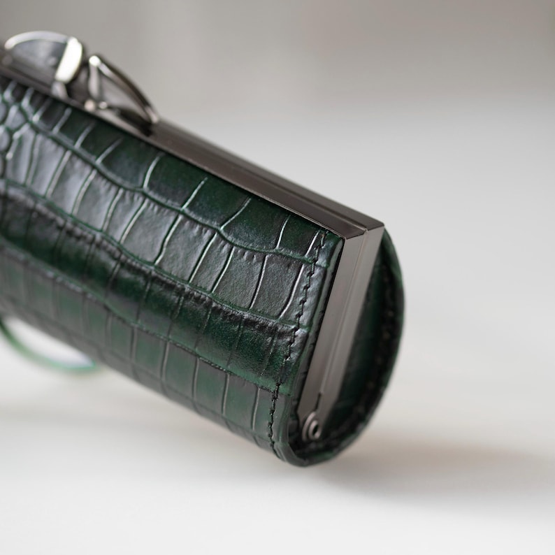 K&ouml;nnte beinhalten: Eine dunkelgr&uuml;ne, rechteckige Clutch mit Krokodilhautstruktur. Die Clutch hat einen dunklen Metallrahmen und -verschluss. Die Tasche hat eine zylindrische Form und scheint aus Leder zu sein.