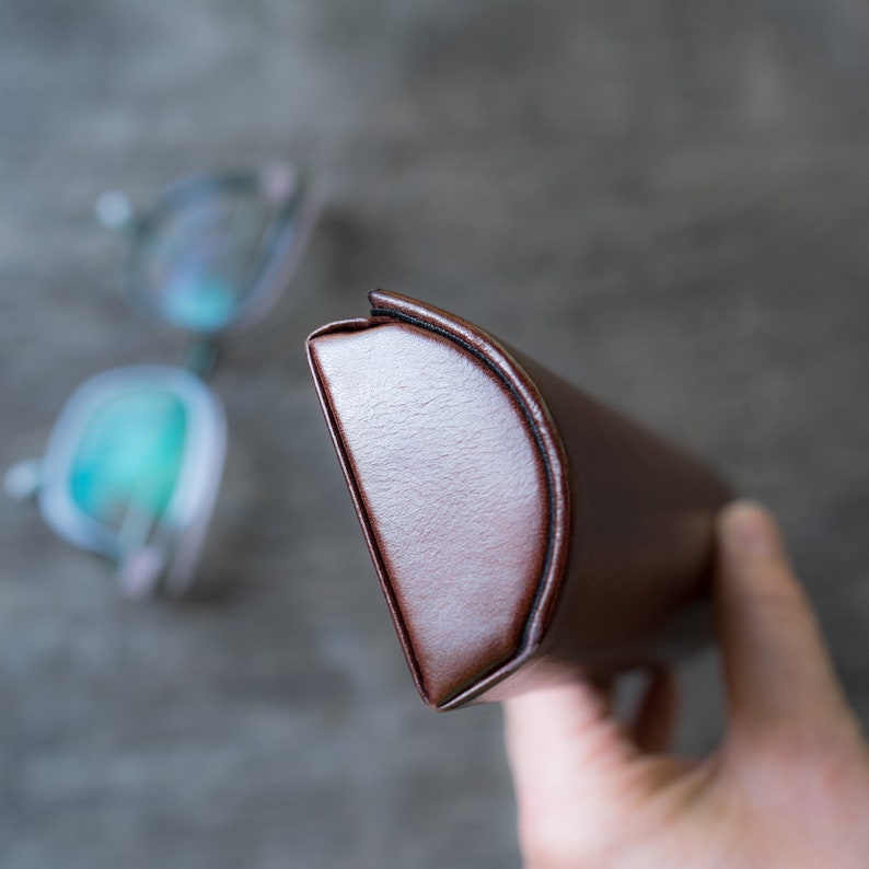 Pu&ograve; includere: Un astuccio per occhiali in pelle marrone con una forma curva. L'astuccio &egrave; chiuso e la parte superiore &egrave; visibile.
