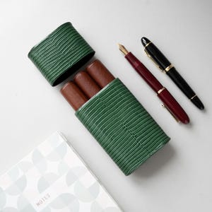 Puede incluir: Un estuche para bolígrafos de color verde texturizado con tapa extraíble, diseñado para tres bolígrafos, con compartimentos interiores marrones. Junto al estuche hay dos plumas estilográficas, una roja oscura con plumín dorado y otra negra con detalles dorados, cerca de un cuaderno.