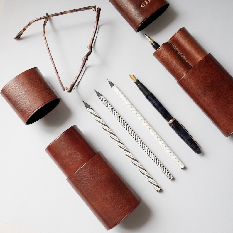 K&ouml;nnte beinhalten: Vier braune Leder-Bleistifthalter, einer mit einem goldenen Stift und vier Bleistiften mit wei&szlig;en und goldenen Streifen.