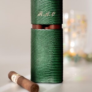 May include: A green, textured leather cigar case with a brown leather band and the initials "R.A.O." in gold. A single cigar rests beside the case. The case is cylindrical and has a reptile-like pattern.