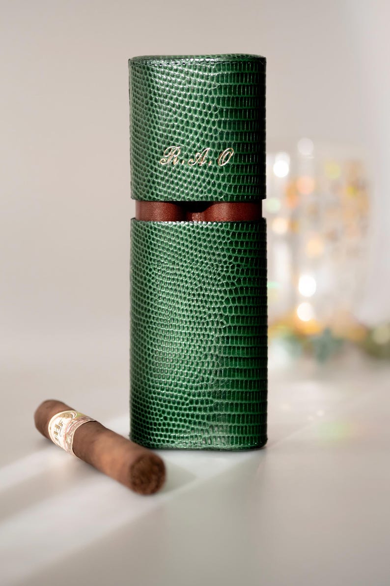 May include: A green, textured leather cigar case with a brown leather band and the initials "R.A.O." in gold. A single cigar rests beside the case. The case is cylindrical and has a reptile-like pattern.