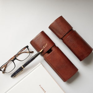 K&ouml;nnte beinhalten: Zwei braune Leder-Stift-Etuis, eines mit den Initialen "CJP" gepr&auml;gt auf der Vorderseite. Ein F&uuml;ller und eine Brille sind ebenfalls abgebildet.