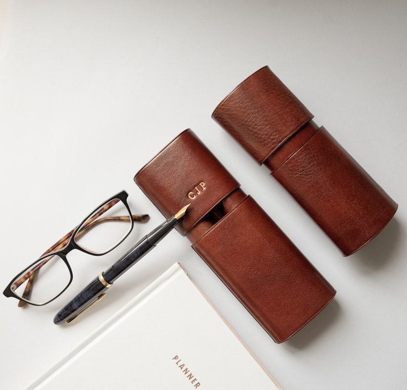 K&ouml;nnte beinhalten: Zwei braune Leder-Stift-Etuis, eines mit den Initialen "CJP" gepr&auml;gt auf der Vorderseite. Ein F&uuml;ller und eine Brille sind ebenfalls abgebildet.