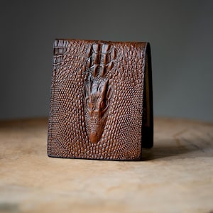 May include: A brown bi-fold wallet featuring a prominent embossed crocodile skin texture across the entire exterior surface. The design showcases a detailed, raised pattern of scales that mimics the appearance of a reptile hide in a rich, dark brown tone.