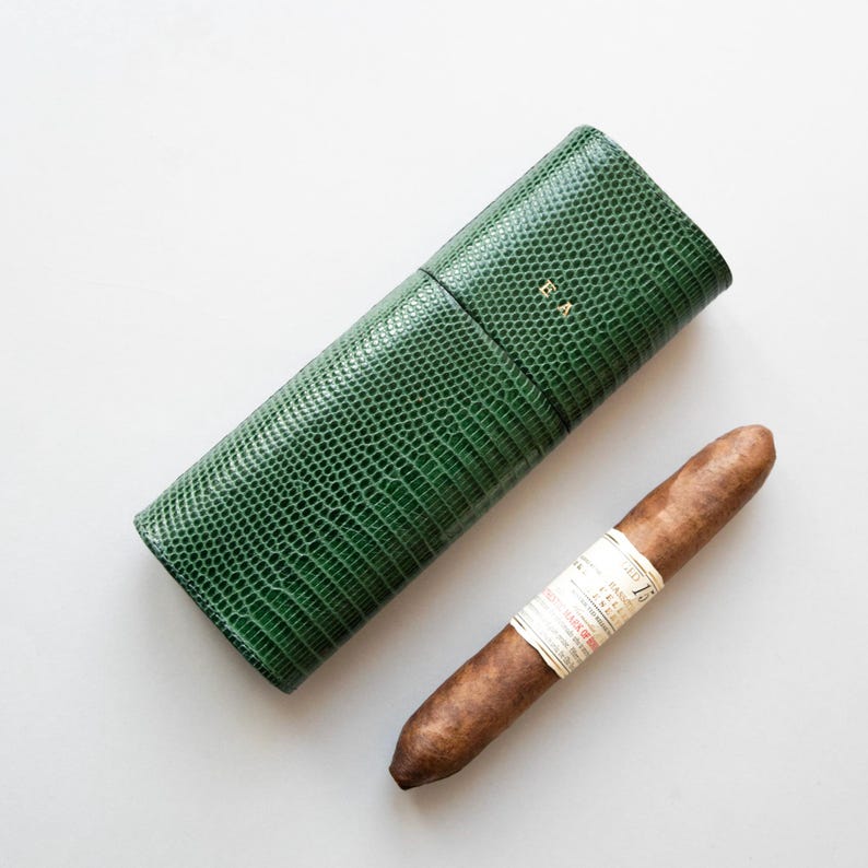 May include: A green, textured cigar case with the initials "E A" in gold lettering, next to a brown cigar with a white label. The case is cylindrical and has a reptile-like pattern.