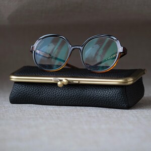 Può includere: Un paio di occhiali con montatura rotonda con dettagli neri e arancioni poggiano su una pochette in pelle nera con chiusura dorata. Gli occhiali hanno lenti trasparenti e aste con motivo tartarugato.