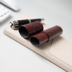 May include: A dark burgundy, crocodile-textured leather pen case consisting of two cylindrical pieces, resting on a beige, pebbled-texture leather mat alongside a black fountain pen with a silver clip and a white star emblem on the cap.